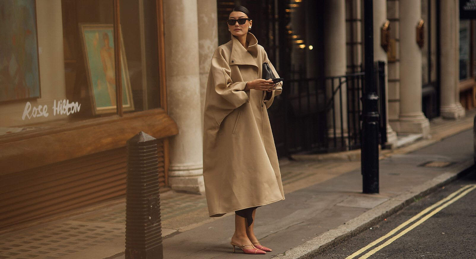 Een modieus geklede vrouw staat op een stoep in een stad, naast een etalage. Ze draagt een oversized, beige trenchcoat met een hoge kraag, en roze puntige muiltjes met een lage hak. Ze houdt een zwarte clutch vast en draagt een zwarte zonnebril. De omgeving bevat klassieke architectuur met pilaren en een etalage met de tekst "Rose Hilton". De sfeer is chic en urban.