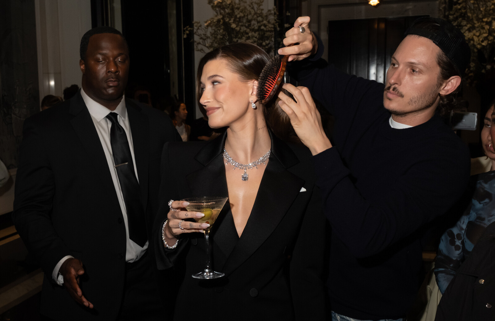 Hailey Bieber bij MET gala met champagne in haar handen en make-up en haarstylisten die haar haar mooi maken lachend in zwarte outfit