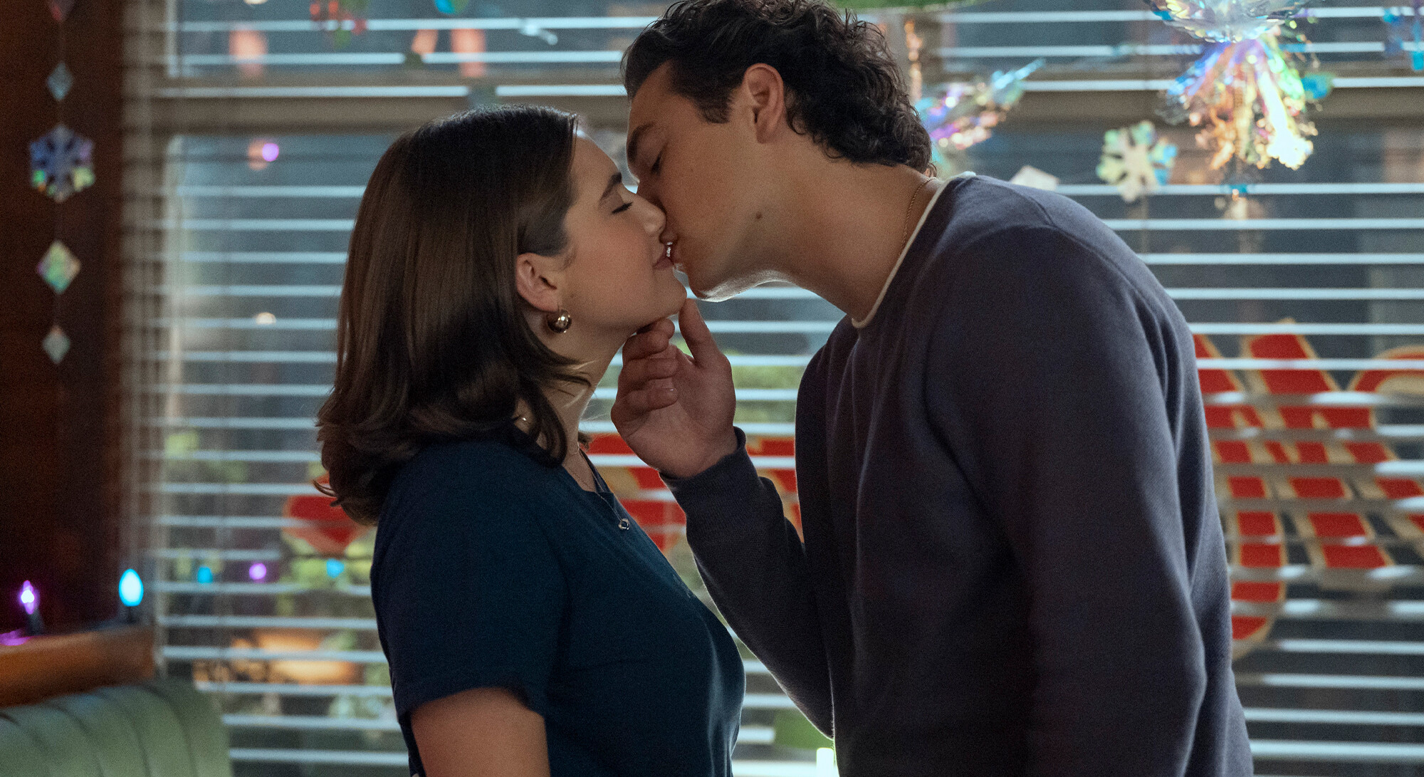 A couple kisses each other in a romantic scene with festive decorations in the background.