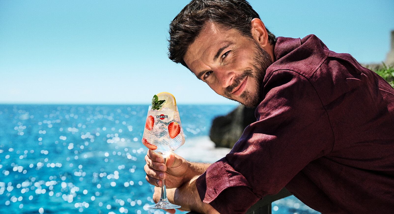 Jonathan Bailey smiling with a Martini cocktail in his hand, summer sea smiling 