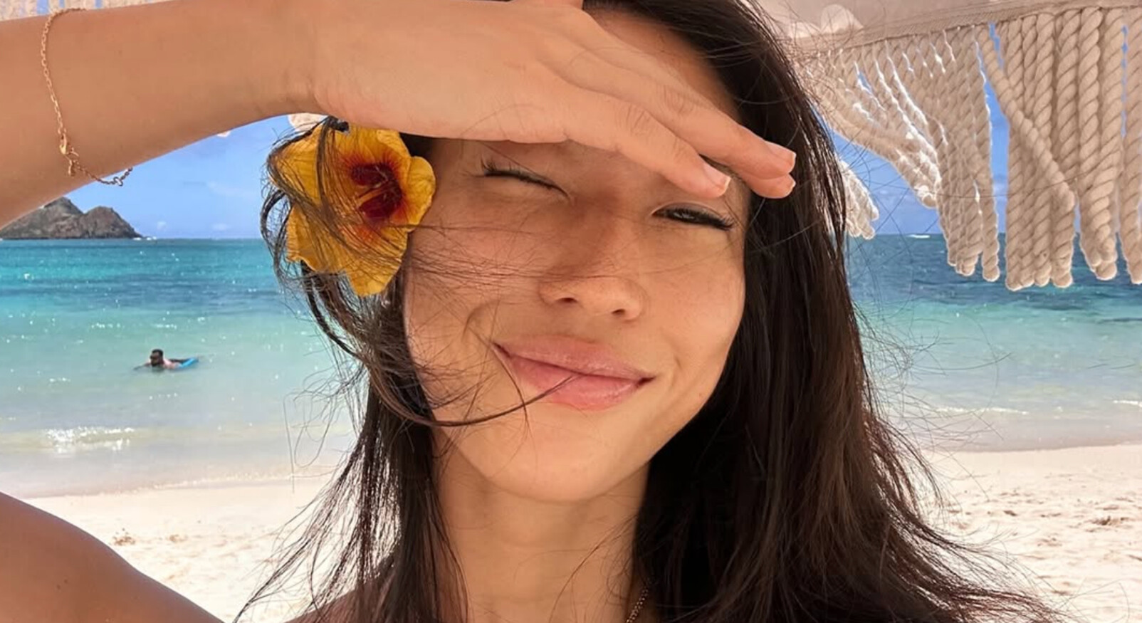 vrouw lachend op het strand met donker haar en bloem achter haar oor zomers bikini lachend