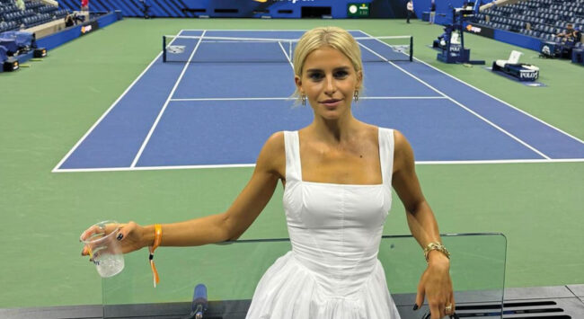 Caro Daur in een witte jurk met een drankje in haar handen lachend bij een tennisbaan, een leegveld