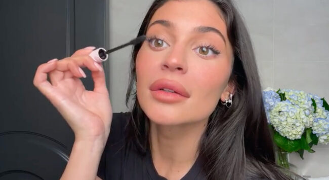 kylie jenner in een make-up video mascara op haar wimpers aanbrengen zwarte top badkamer bloemen op de achtergrond