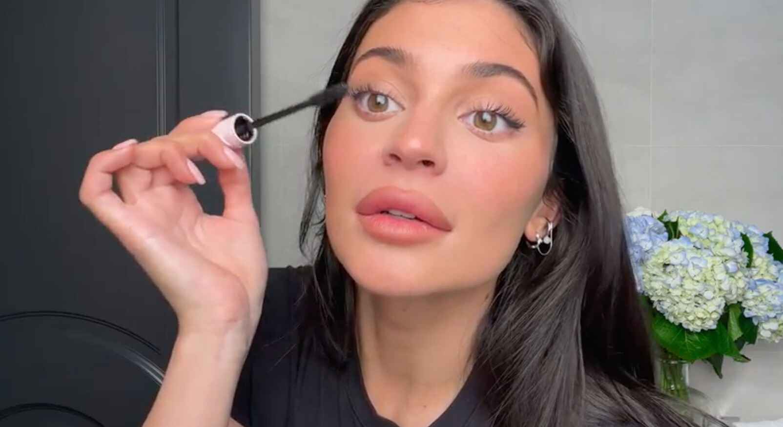 kylie jenner in een make-up video mascara op haar wimpers aanbrengen zwarte top badkamer bloemen op de achtergrond