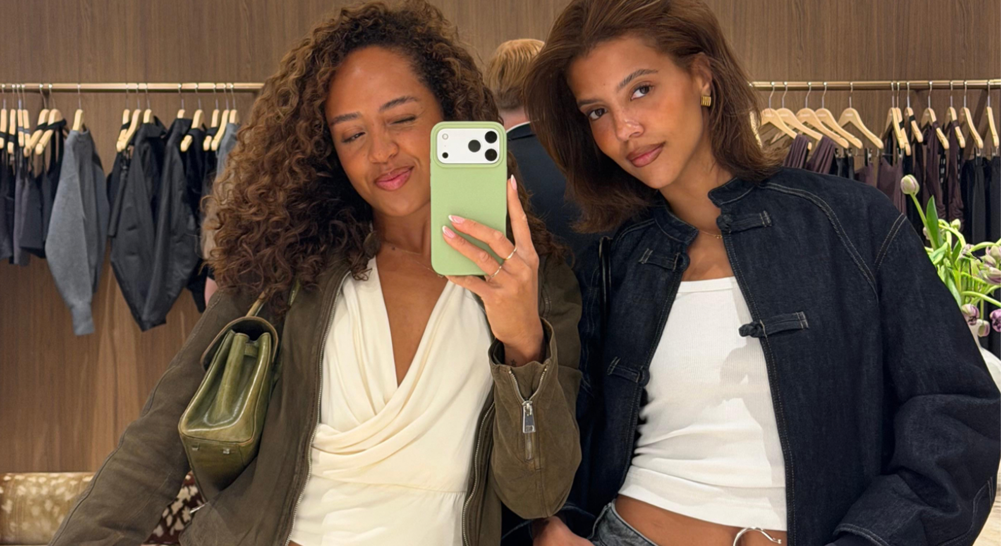 Twee vrouwen maken een spiegelselfie in een kledingwinkel, met volumineus haar en een frisse beautylook.