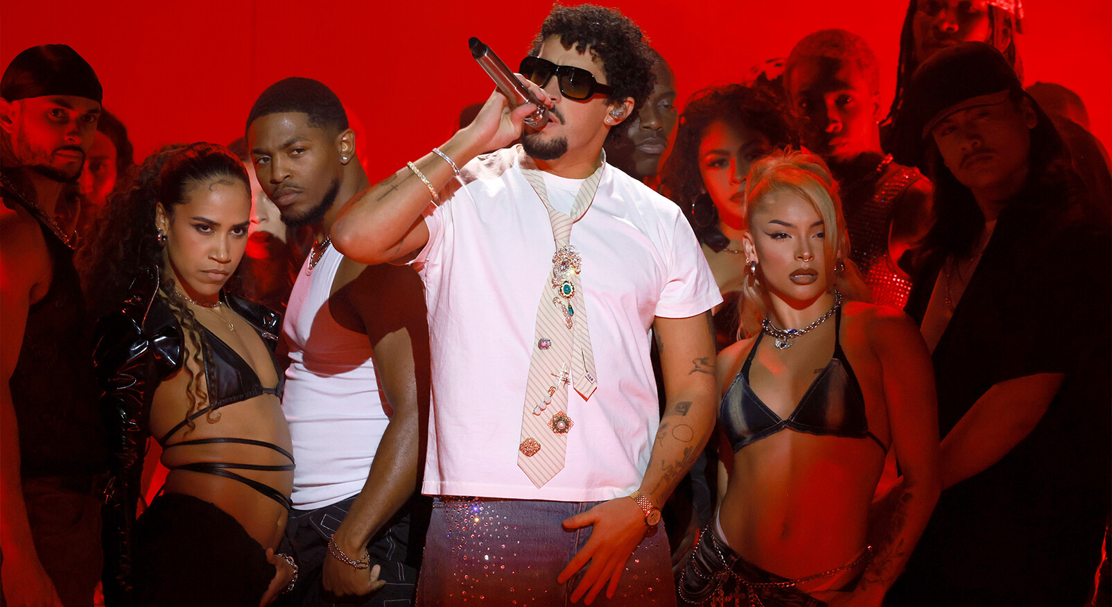 De Puerto Ricaanse reggaeton- en latin trap-artiest Bad Bunny staat op een podium, omringd door achtergronddansers, tijdens een optreden. Hij draagt een wit T-shirt, een zonnebril, een bejewelde stropdas en een glinsterende spijkerbroek terwijl hij een microfoon vasthoudt. De achtergrond is verlicht in een intense rode kleu