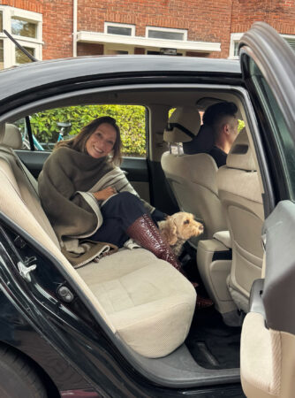 A woman is sitting in the back seat of a black car and is smiling at the camera. She is wrapped in a warm, olive green blanket and is wearing a shiny, burgundy boot on the seat. Next to her is a small, light brown, curly-haired dog, looking out the window. The background is a brick wall of a house.
