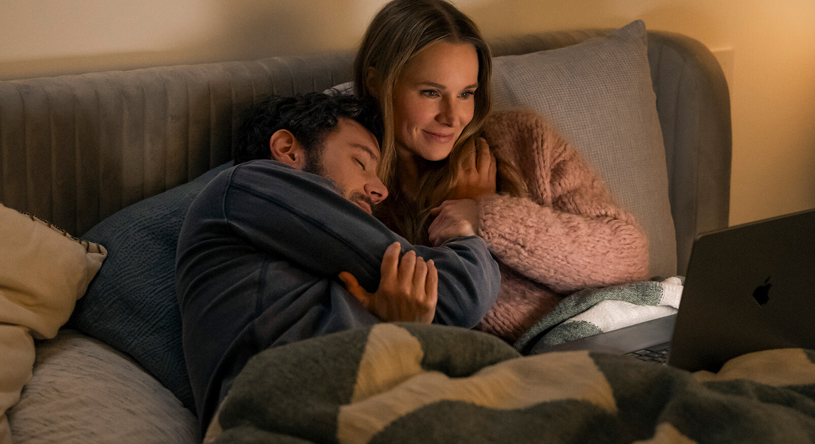 Een man en vrouw liggen knus tegen elkaar aan in bed, met een deken over hen heen, en kijken samen naar een laptop met een Apple-logo dat op hun schoot staat. De man, met een baardje, ligt met zijn hoofd tegen de borst van de vrouw aan, en heeft zijn armen om haar heen geslagen. De vrouw kijkt met een zachte glimlach naar het scherm en draagt een roze, harige trui. De slaapkamer is warm verlicht.