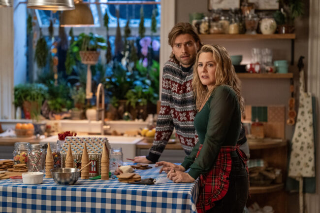 Een man en een vrouw staan achter een tafel in een gezellige, goed verlichte keuken. De vrouw, met blond haar en gekleed in een donkergroene trui en een rood-geruite schort, leunt licht naar voren en kijkt met een verraste of verontruste uitdrukking. De man, met langer blond haar en gekleed in een Noorse kersttrui met rood en wit patroon, staat iets achter haar en heeft dezelfde verbaasde blik. Op de blauw-wit geruite tafel liggen verspreid koekjes, kleine chocoladesnoepjes, en er staan zilveren kommen en wafelkegels, wat suggereert dat ze bezig waren met bakken of het versieren van peperkoekhuisjes. Op de achtergrond is een gootsteen en een groot raam met planten te zien, en aan de rechterkant zijn open planken gevuld met potten en etenswaren."