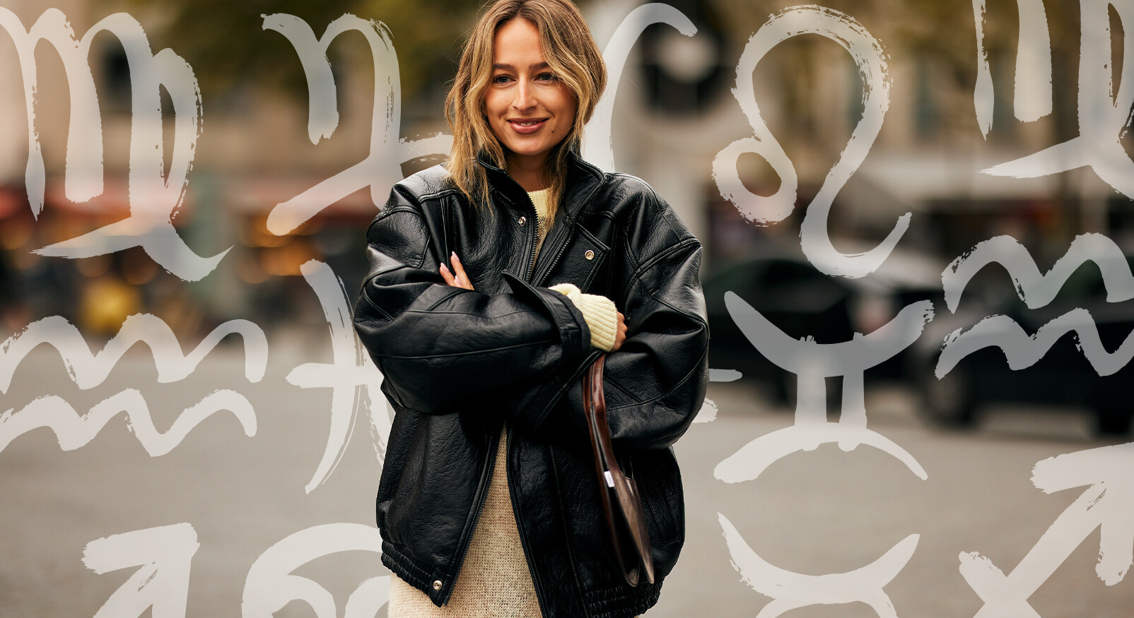 Een modieuze vrouw met halflang blond haar staat met de armen over elkaar in het midden van de foto. Ze draagt een oversized, zwarte leren jas over een lichtgeel kledingstuk. De achtergrond is onscherp met grijze en bruine tinten, en er zijn transparante witte/grijze astrologische tekens van de dierenriem over de hele afbeelding heen gelegd.