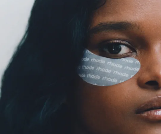 Een close-up van het gezicht van een vrouw, haar rechteroog is open. Onder haar oog is een donkergrijs oogmasker (eye patch) geplakt, met het woord 'rhode' herhaald in witte letters. Haar donkere haar valt langs haar wang.