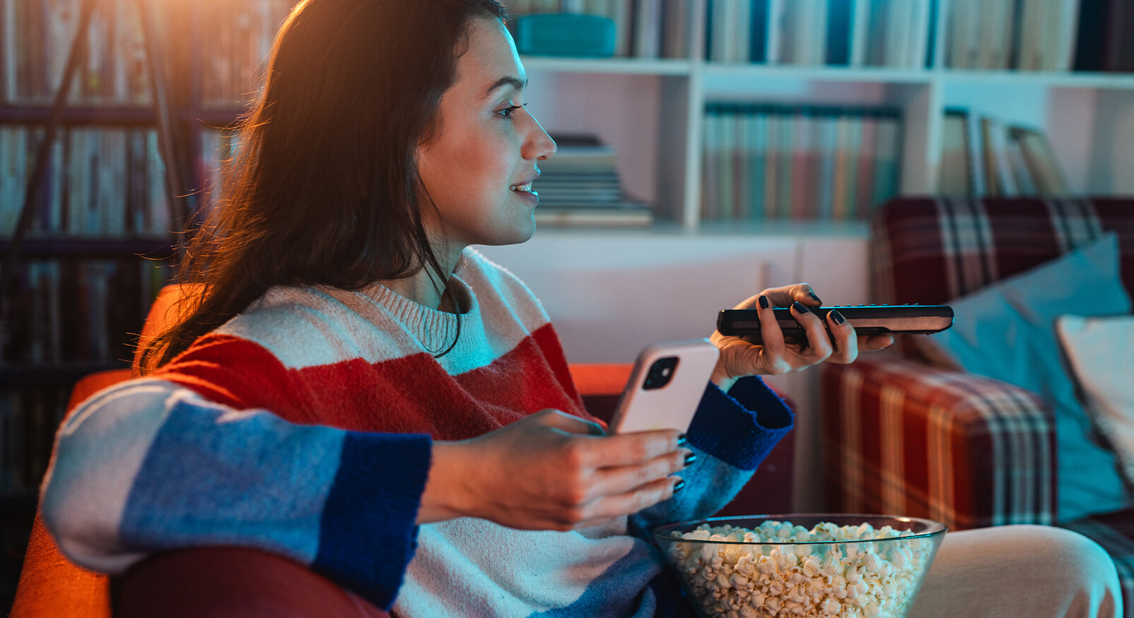 A close-up of a smiling young woman with long, dark hair sitting on a couch in a warmly lit living room. She is wearing a red-white-blue sweater and looking sideways at a screen (not visible). In her left hand, she holds a white smartphone and in her right hand a remote control. On her lap is a large, transparent bowl of popcorn. In the background, bookshelves are visible.