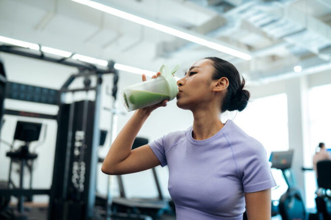 Een vrouw in sportkleding met opgestoken haar drinkt uit een shakerfles met een groene vloeistof in een sportschool. Ze draagt een lichtpaars T-shirt. Fitnessapparatuur en een andere persoon zijn wazig op de achtergrond te zien.