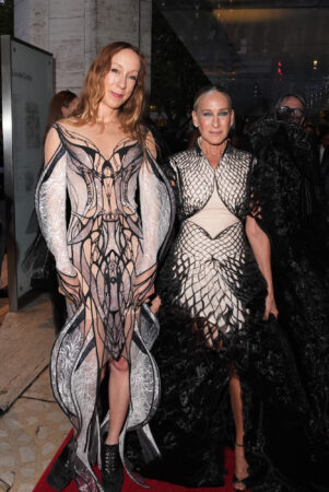 Een foto van de Amerikaanse actrice Sarah Jessica Parker (rechts) en modeontwerper Iris van Herpen (links) die naast elkaar op een rode loper staan. Sarah Jessica Parker draagt een crèmekleurige jurk met een ingewikkelde, zwarte, schubachtige overlay en een grote zwarte cape van texturen. Iris van Herpen draagt een van haar kenmerkende ontwerpen: een witte en lichtroze jurk met complexe, donkere, abstracte lijnen en uitsnijdingen die doen denken aan vleugels of een skelet. Beide vrouwen kijken recht in de camera.