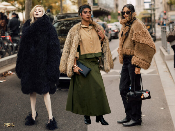 Drie vrouwen in opvallende jassen met bont- of shaggy textuur. Links een vrouw in een dramatische, lange zwarte, harige jas met gevederde zwarte schoenen. Midden een vrouw in een beige/tan corduroy overhemd en een lange donkergroene rok (passend bij jouw favoriete kleur groen) met een beige bontjas. Rechts een vrouw in een cropped, shaggy bruine jas en zwarte jeans.