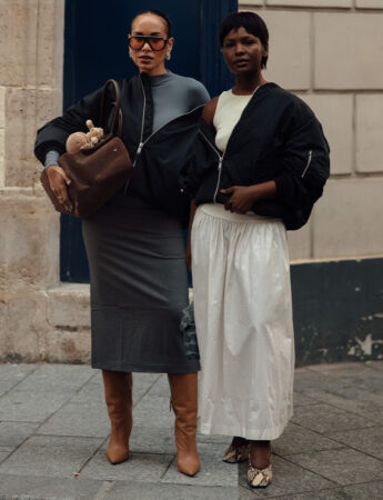 "Twee vrouwen in casual-chique outfits met oversized zwarte bomberjacks. De vrouw links draagt een grijze kokerrok, een grijs nauwsluitend topje en hoge bruine leren laarzen. De vrouw rechts combineert een wit topje en een wijde witte lange rok met lage hakken met slangenprint."