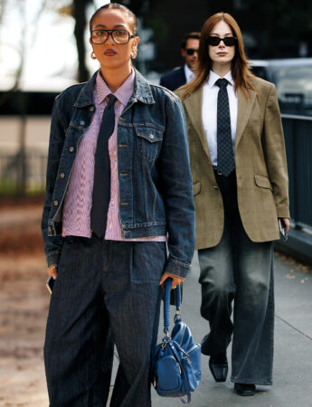 "Twee vrouwen met een verfijnde streetstyle in pakken en wijde broeken. De vrouw links draagt een gestreept overhemd en stropdas onder een denim jack en een wijde spijkerbroek, met een felblauwe handtas. De vrouw rechts draagt een bruine blazer, een witte blouse, een stropdas en een wijde, donkergewassen spijkerbroek."