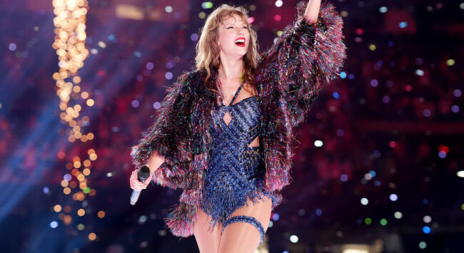 Taylor Swift treedt op tijdens de Eras Tour, gekleed in een glinsterende donkerblauwe bodysuit met cut-outs en een kleurrijk, harig, kort jasje in blauwe, roze en paarse tinten. Ze staat op het podium met haar rechterarm omhoog, terwijl confetti over de menigte valt. Haar haar is los en golvend, en ze draagt felrode lippenstift.