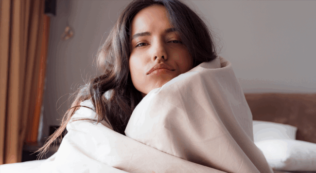 Vrouw met donkere haren zit in bed, gewikkeld in een deken met een ongemakkelijke blik, symbolisch voor bijwerkingen.