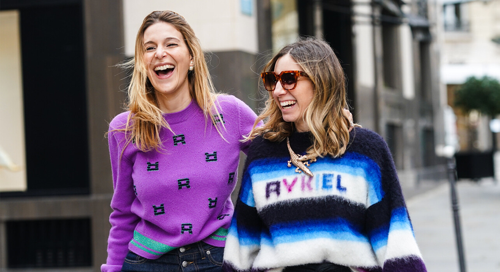 Twee vrouwen in opvallende truien (paars en blauw/wit) lachen uitbundig op straat, symbool voor humor en grappige sterrenbeelden.