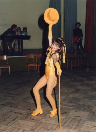 "Victoria Beckham als jong meisje, waarschijnlijk in 1986, in een gele dansvoorstelling outfit. Ze staat op een houten podium, houdt een gele bolhoed met gestrekte arm omhoog en leunt op een wandelstok, met een brede glimlach. Op de achtergrond zitten enkele mensen aan tafels."