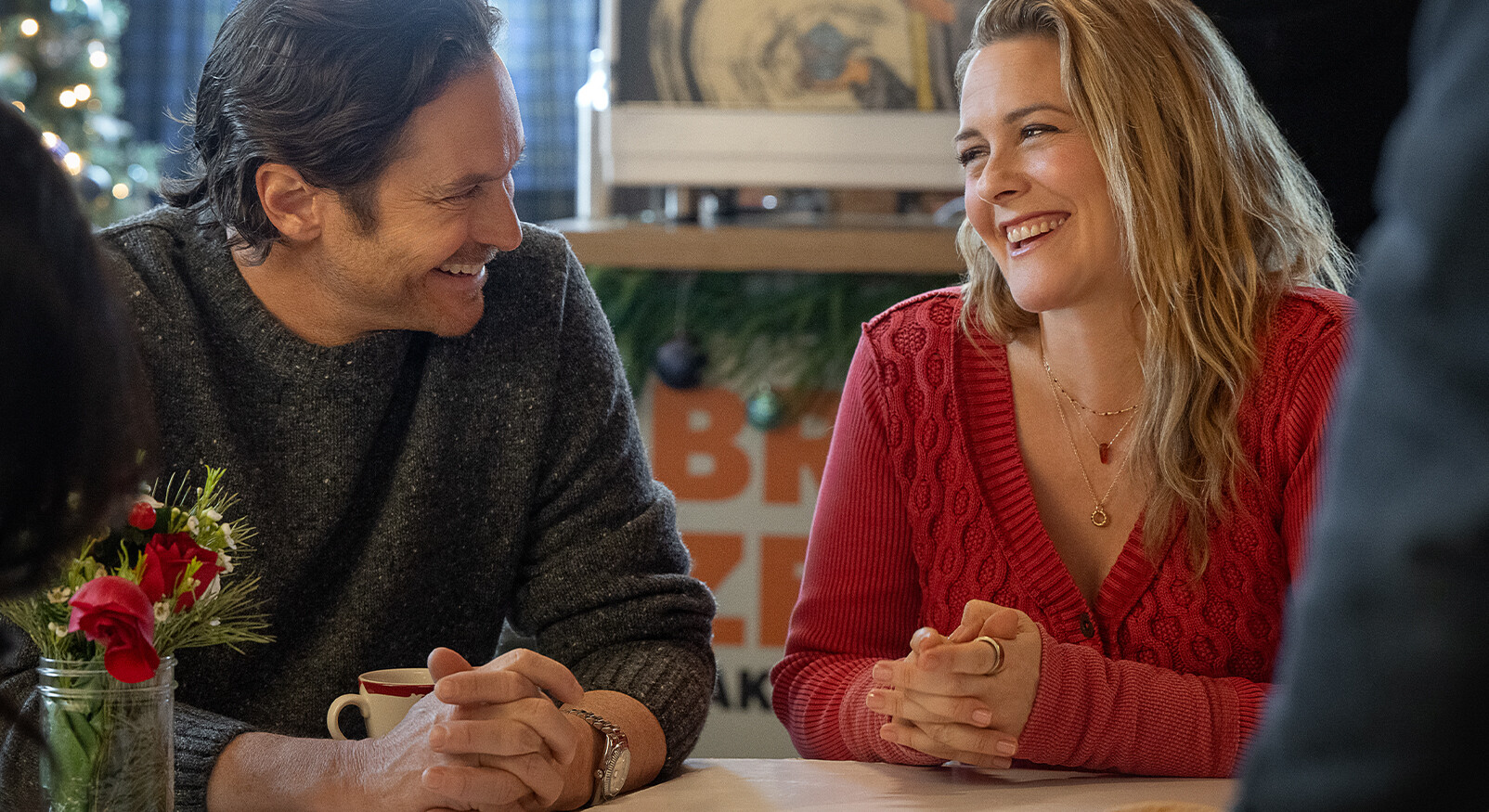 "Een close-up van een lachende man en vrouw die tegenover elkaar zitten aan een tafel, mogelijk in een café of restaurant. De vrouw, met blond haar en een rode gebreide trui, kijkt de man met een brede lach aan. De man, met donker haar en een donkergrijze, grofgebreide trui, lacht ook en kijkt naar de vrouw. Ze hebben allebei hun handen op tafel gevouwen. Op de voorgrond is een glazen pot met rode bloemen te zien en op de achtergrond een onscherp kerstdecor."