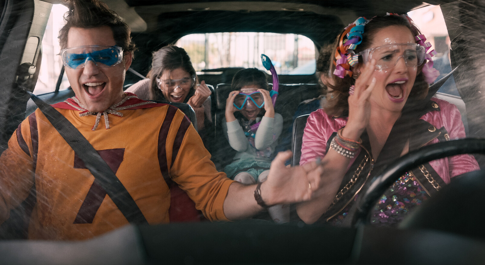 "Een gezin van vier, gekleed in kostuums of met accessoires zoals duikbrillen en snorkels, zit in een auto. De vader, gekleed als superheld in een geel-rood kostuum met cape en blauwe skibril, en de moeder (met roze jasje en haarlinten) schreeuwen of zingen enthousiast. De twee kinderen op de achterbank lachen of kijken verbaasd. Het raam is bespat, wat de chaos en avontuurlijke sfeer benadrukt."