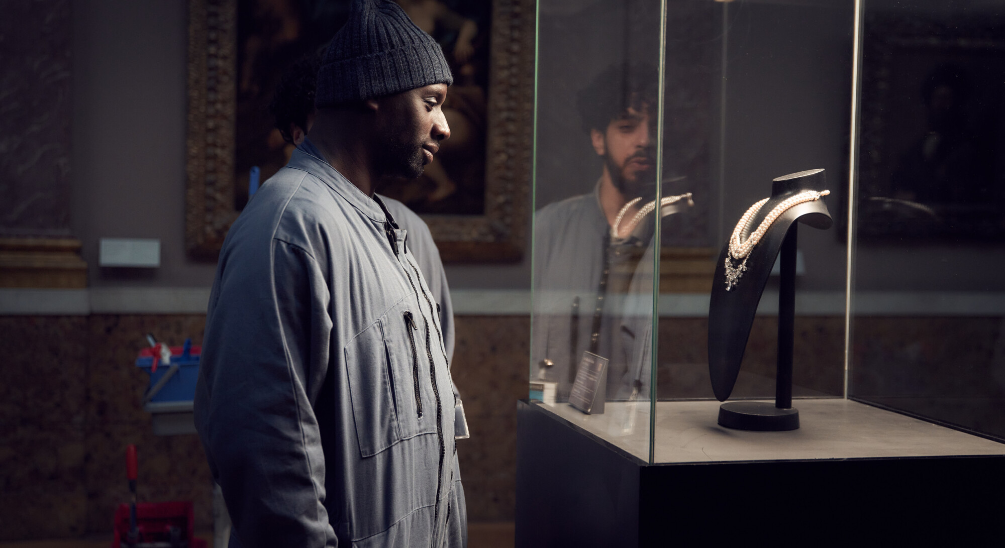 Een man in een grijs overall en een donkerblauwe muts bekijkt een diamanten halsketting die in een glazen vitrine ligt in een museum. Achter het glas is ook een andere man te zien die zijn reflectie spiegelt.