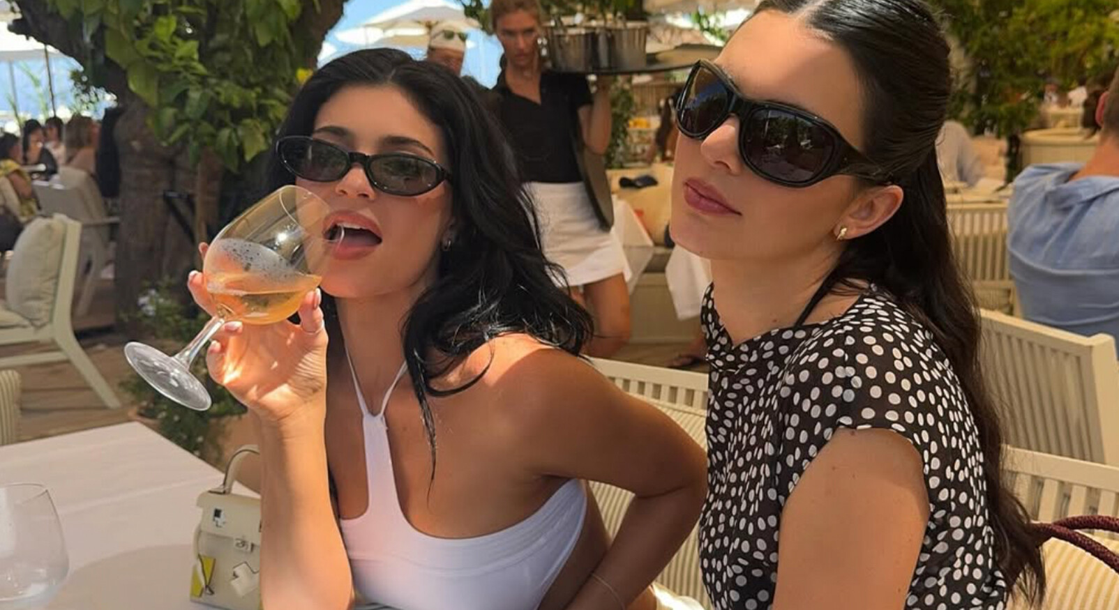 Twee vrouwen zitten aan een tafel buiten op een zonnig terras. De vrouw links, met lang donker haar en een witte, getailleerde top, draagt een donkere zonnebril en drinkt uit een glas met een oranjeroze drankje. De vrouw rechts heeft ook lang donker haar, draagt een donkere zonnebril en een zwart-wit gestippelde top. Op de achtergrond is een ober in een wit overhemd met een dienblad te zien en er staan bomen en andere tafels. De sfeer is luxe en zomers.