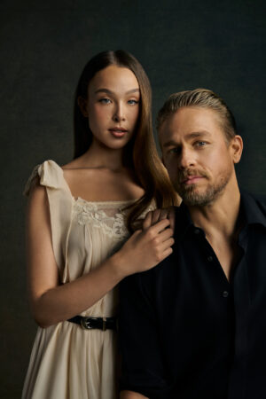 Alt-text for "MONSTER_S4_GLAM_CHARLIE_ELLA_Seamless_00257_FINAL.jpg":  A formal, dark-toned promotional photo for the 'Monster' season about Lizzie Borden. The photo shows actress Ella Beatty (who plays Lizzie Borden) and actor Charlie Hunnam (who plays Andrew Borden). Ella Beatty stands on the left in a light, cream-colored 19th-century style dress with lace details and a black belt, and has her hand on Charlie Hunnam's shoulder. Charlie Hunnam stands on the right in a dark, open shirt with his hair slicked back. Both look seriously at the camera.