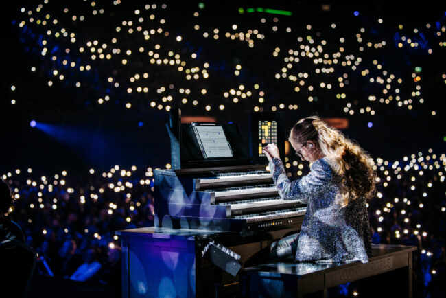 "A young woman, seen from behind, with long wavy hair and a sparkling silver jacket, is concentrating behind a large digital organ with multiple keyboards and sheet music. She is performing for a large, dark hall, whose audience is visible through the thousands of small, bright lights of mobile phones or flashlights held above their heads."