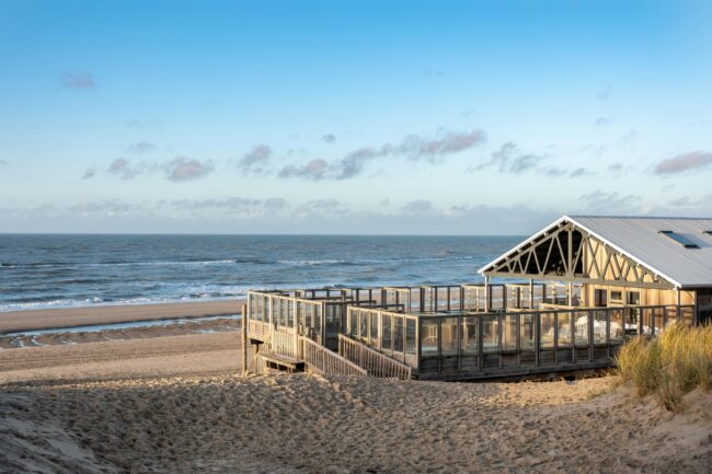 A wide, horizontal landscape of a beach pavilion by the sea on a sunny day. The pavilion is a wooden structure with a gray gabled roof, partly built on an elevated platform. The platform has a wooden railing with glass panels and is accessible via a wooden staircase that runs up from the beach. The pavilion stands on the wide sandy beach, with dune grass on the right in the foreground. The sea stretches out to the horizon under a clear blue sky with a few light clouds.