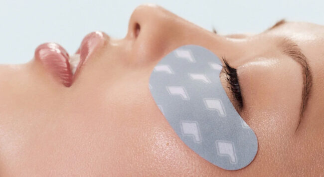 Een close-up van een vrouw die op haar rug ligt met haar ogen gesloten. Op haar wang en onder haar gesloten oog ligt een lichtblauw/grijs oogmasker (eye patch) met een herhalend ruitpatroon in wit. Haar huid ziet er glanzend en goed gehydrateerd uit.