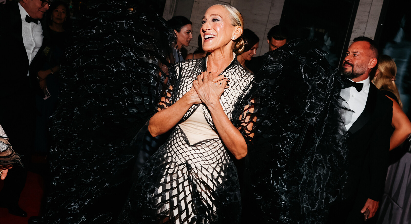 Een foto van de Amerikaanse actrice Sarah Jessica Parker op een rode loper, mogelijk tijdens een gala. Ze draagt een opvallende jurk met een lichte, korset-achtige top en een lange, wijd uitwaaierende rok en cape van zwart, textuurrijk materiaal dat er vederachtig of geplooid uitziet. Ze heeft haar handen over haar borst gevouwen en lacht breed en opgetogen. Op de achtergrond zijn andere, formeel geklede gasten te zien en een verlichte muur met tekst.