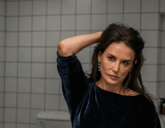 Een medium shot van een vrouw in een donkerblauwe fluwelen top die voor een spiegel staat in wat een badkamer lijkt te zijn, met witte vierkante tegels op de muur. Ze kijkt recht in de camera terwijl ze haar hand in haar donkere haar houdt.