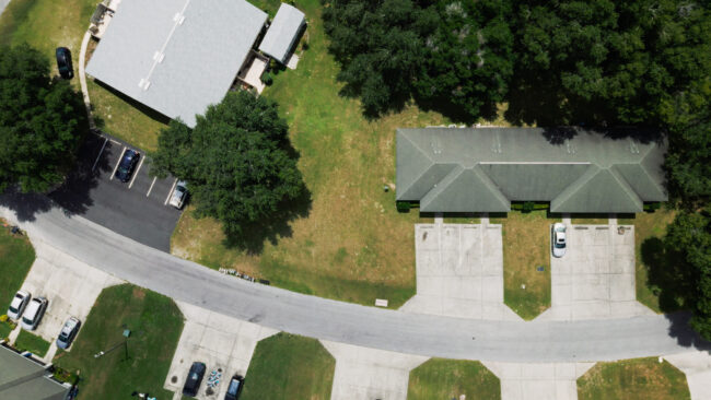 Luchtfoto genomen vanuit vogelperspectief van een woonwijk met groene grasvelden en bomen. De afbeelding toont een tweevoudig appartementengebouw met een grijsgroen dak en een parkeerplaats ervoor met één witte auto. Links daarvan staat een vierkant, lichtgekleurd gebouw met een parkeerplaats. Een gebogen weg loopt door het midden.