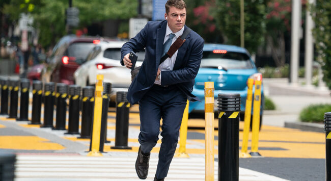 A man in a suit running across a street, holding a phone and looking determined, with cars in the background.