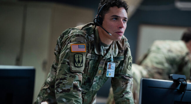 A soldier in military uniform, wearing a headset and focused on a task in a high-tech control room.