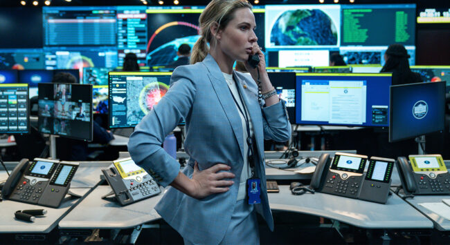 A woman in a blue suit standing in a control room, speaking on the phone while surrounded by multiple computer screens and telephones.