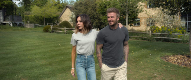 Victoria en David Beckham lopen in de zon door een grote, groene tuin. Victoria draagt een lichtgrijs T-shirt en een high-waist spijkerbroek, David draagt een donkergrijs T-shirt en beige broek. Op de achtergrond staat een landelijk huis met een rieten dak en een houten hek."