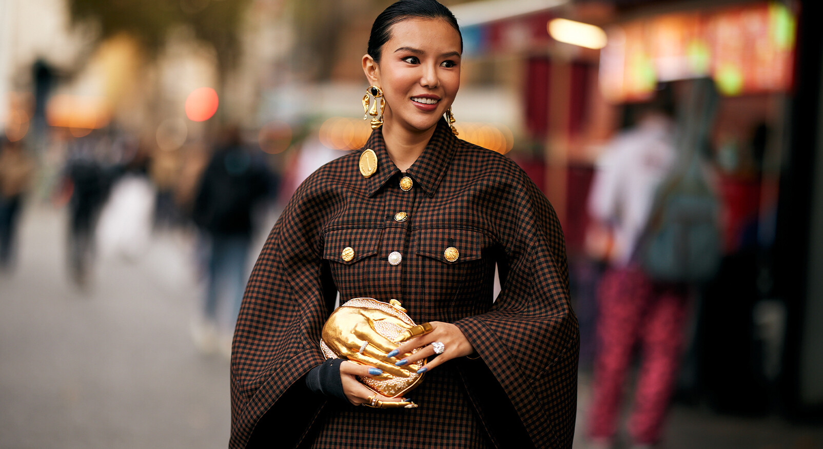 "A stylish woman with her hair slicked back poses on a busy street. She wears a brown-black checkered cape with large gold buttons and striking, sculptural gold earrings. She holds a unique, gold clutch in the shape of a hand with bright blue nail polish. The filename links the image to the theme of zodiac signs and fetishes."
