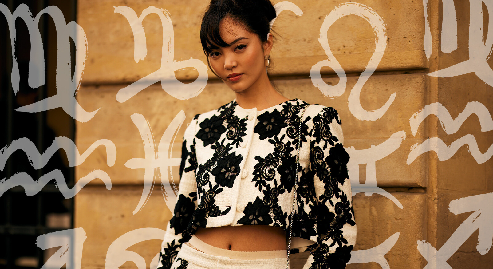 Een modieuze vrouw staat buiten en kijkt recht in de camera. Ze draagt een zwart-witte, getailleerde crop-top met een gestructureerd, gebloemd kantpatroon. Haar haar is opgestoken en ze draagt subtiele gouden oorbellen. De achtergrond is een beige, stenen gebouw, en de afbeelding is gelaagd met witte, abstracte illustraties van astrologische tekens (zoals Steenbok, Vissen, Leeuw en Schorpioen) die het thema van een horoscoop suggereren.