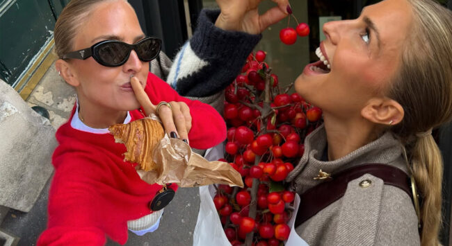 Two blonde women smile and enjoy snacks outside. The woman on the left, dressed in a bright red jumper and black sunglasses, holds a croissant and brings her finger to her lips in a 'still' gesture. The woman on the right wears a beige jacket and smiles while holding a red cherry she has picked from a large bunch of shiny, bright red berries or small apples in the centre of the picture.