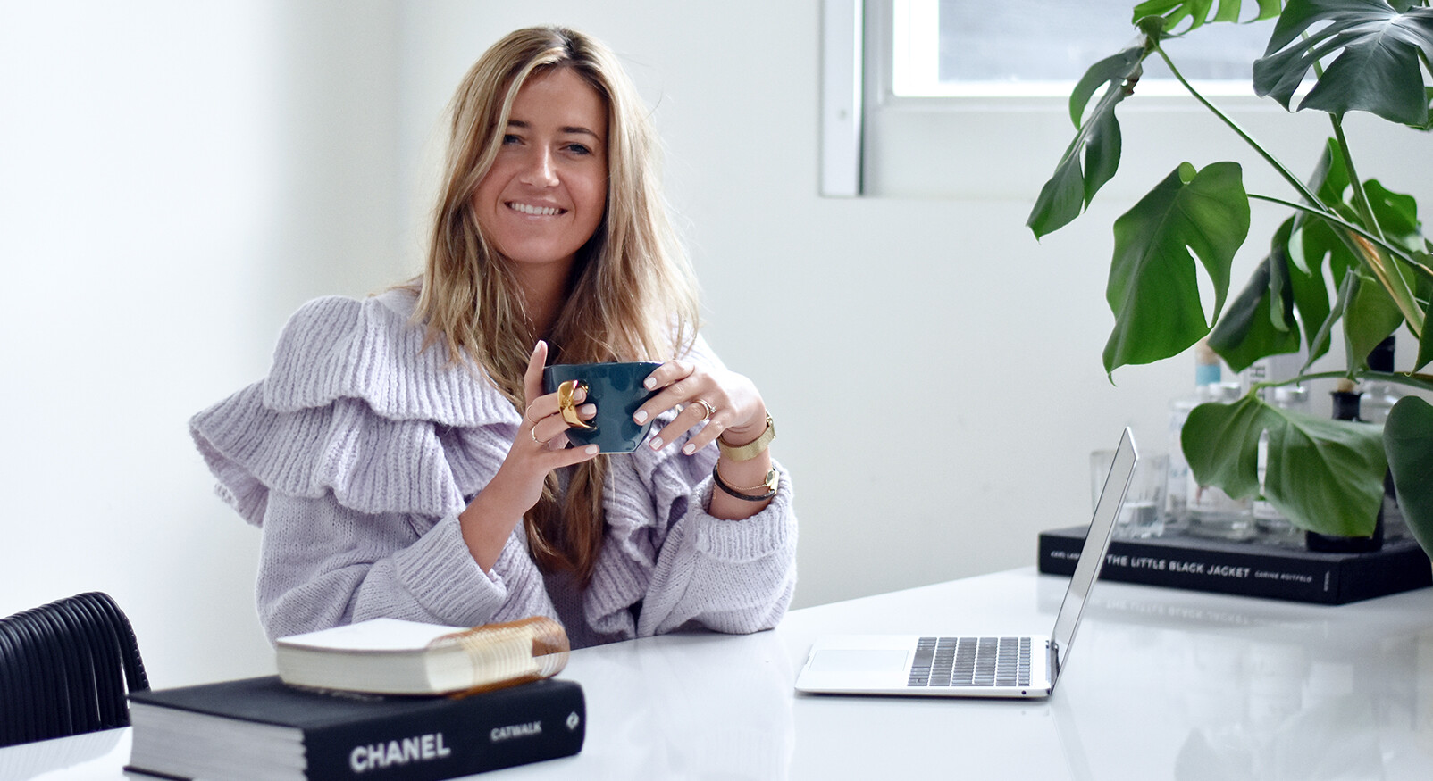 Een lachende, blonde vrouw in een lichtpaarse trui met ruches zit aan een witte tafel. Ze houdt een donkergroene mok vast. Voor haar staat een opengeklapte zilveren laptop en een stapel boeken, waaronder één met 'CHANEL' erop. Rechts staat een grote, groene kamerplant. De achtergrond is wit, met een raam dat zacht licht binnenlaat.