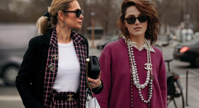 Twee modieuze vrouwen lopen buiten in een stadse omgeving. De vrouw links draagt een zwart-roze tweed blazer, een witte top en een zwarte zonnebril. De vrouw rechts draagt een paarse trui over een witte blouse, meerdere opvallende parelkettingen en een zwarte zonnebril. Beide vrouwen hebben een elegante, chique uitstraling.