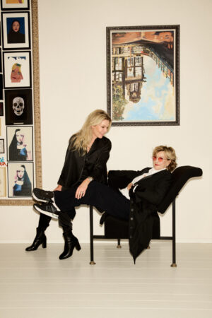 Two blonde women pose in a modern room with art on the wall. A younger woman, dressed in a black satin blouse, dark pants, and high black boots, sits on the arm of a black chaise longue. An older woman, with a short blonde haircut and pink-framed glasses, lies in the chaise longue wearing a black blazer over a white shirt. Behind them hangs a large painting of an Amsterdam canal and a collage of smaller, framed artworks.