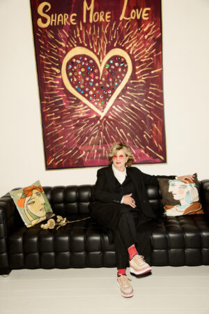 An older blonde woman sits relaxed on a black leather Chesterfield-style sofa. She wears a black pinstripe suit, a white shirt, pink/red socks, and striking beige-pink sneakers with a thick sole. She looks straight into the camera with pink-framed glasses. On the sofa are two cushions with illustrations of faces. Behind her hangs a large painting with the text 'Share More Love' above a golden heart, surrounded by golden rays on a burgundy background. There is also a bunch of golden flowers on the sofa.