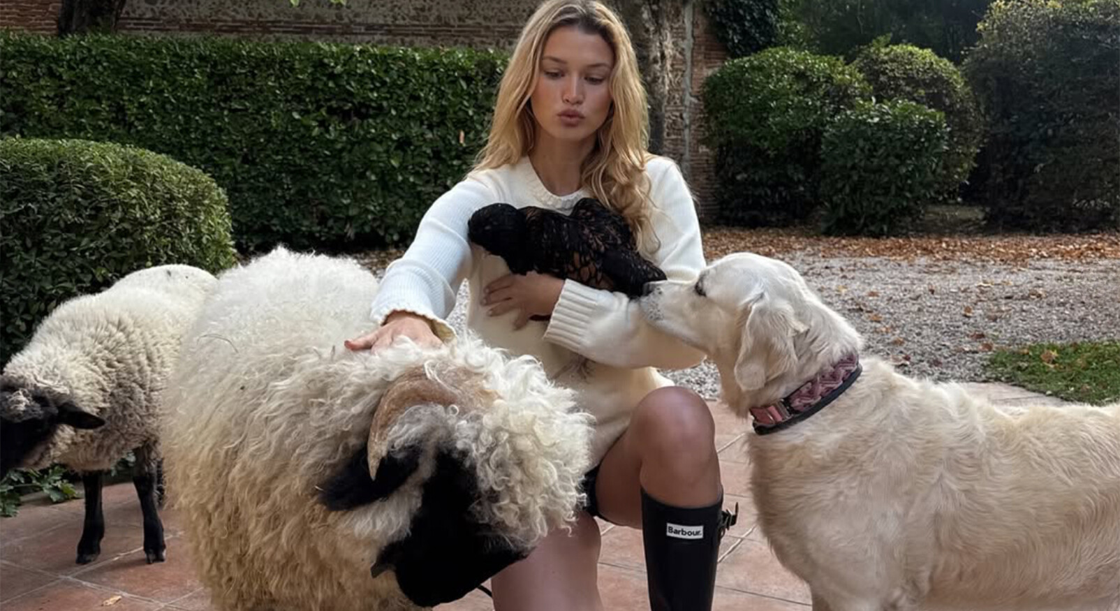 Een lichte, jonge vrouw met lang blond haar, zittend op een terras buiten, draagt een crèmekleurige gebreide trui en zwarte regenlaarzen. Ze wordt omringd door dieren: ze houdt een klein, zwart kipje tegen haar borst en aait een groot, wollig schaap met zwarte kop, dat links van haar zit. Rechts van haar is een blonde golden retriever te zien die haar schaap of haar gezicht snuffelt. Op de achtergrond is een groene haag en een bestraat terras te zien.