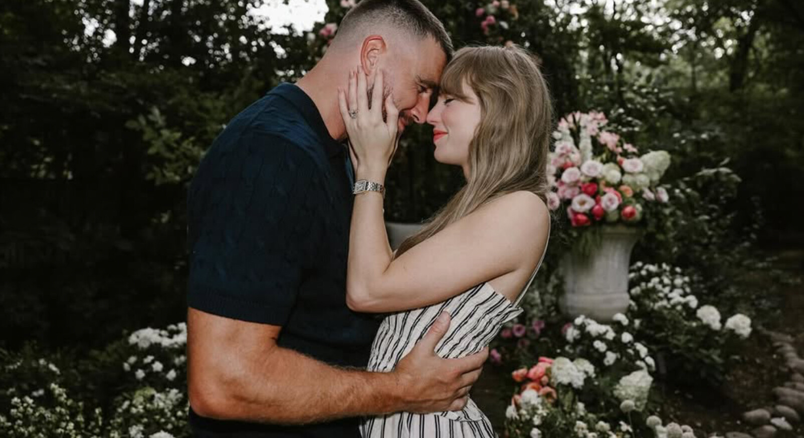 Een verliefd stel staat dicht tegen elkaar in een weelderige tuin vol bloemen. De vrouw houdt het gezicht van de man teder vast terwijl ze met hun voorhoofden tegen elkaar leunen. Op de achtergrond staat een grote vaas met kleurrijke bloemen tussen het groen.