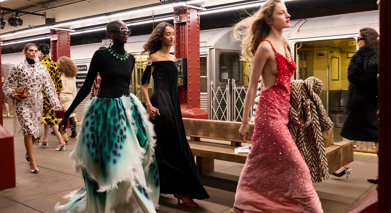 Zes modellen lopen langs een houten bank op een ondergronds metrostation in New York. De modellen dragen de Chanel Pre-Fall collectie, variërend van een lichtblauwe jeans/shirt look en een wijde blauwe jurk tot een man in een spijkerbroek en bomberjack, en een model in een zwarte outfit met een opvallende rode tailleband. Een metrostel staat stil op de achtergrond en de pilaren zijn rood.